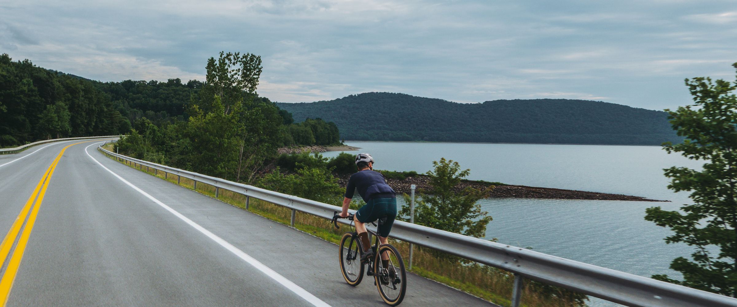 A lone rider on an empty road above a reservoir, forested ridge and weather beyond.