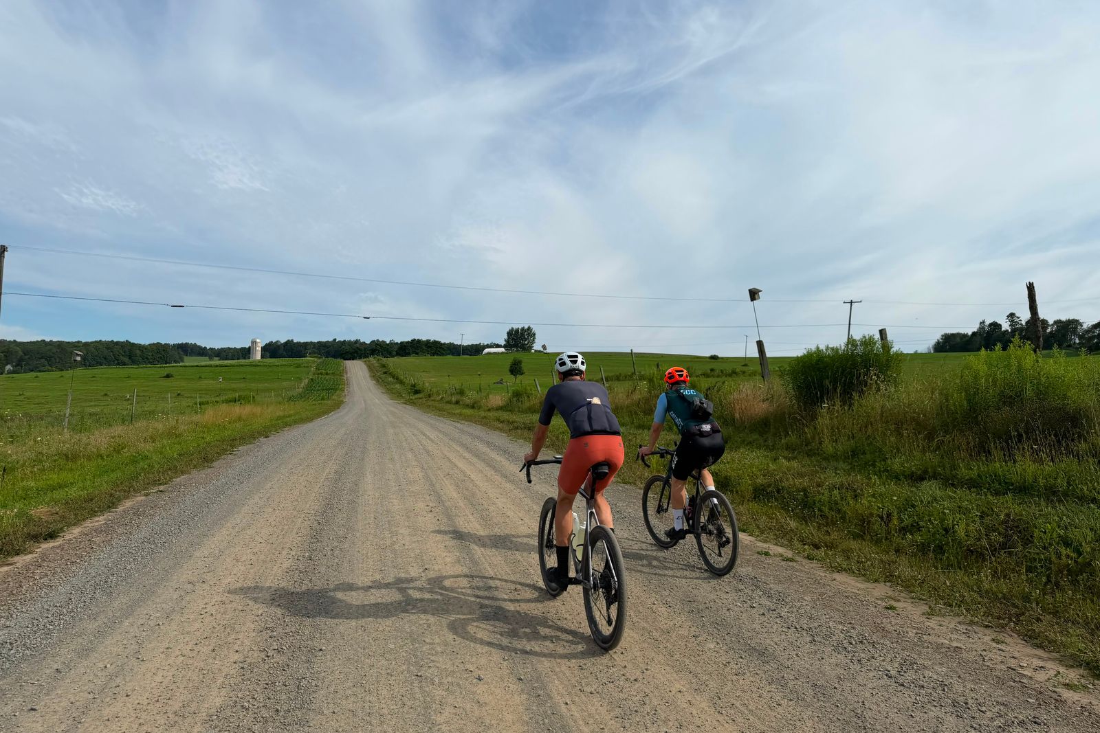 Two riders on a gravel road running out through open pasture toward a distant silo, big sky above.