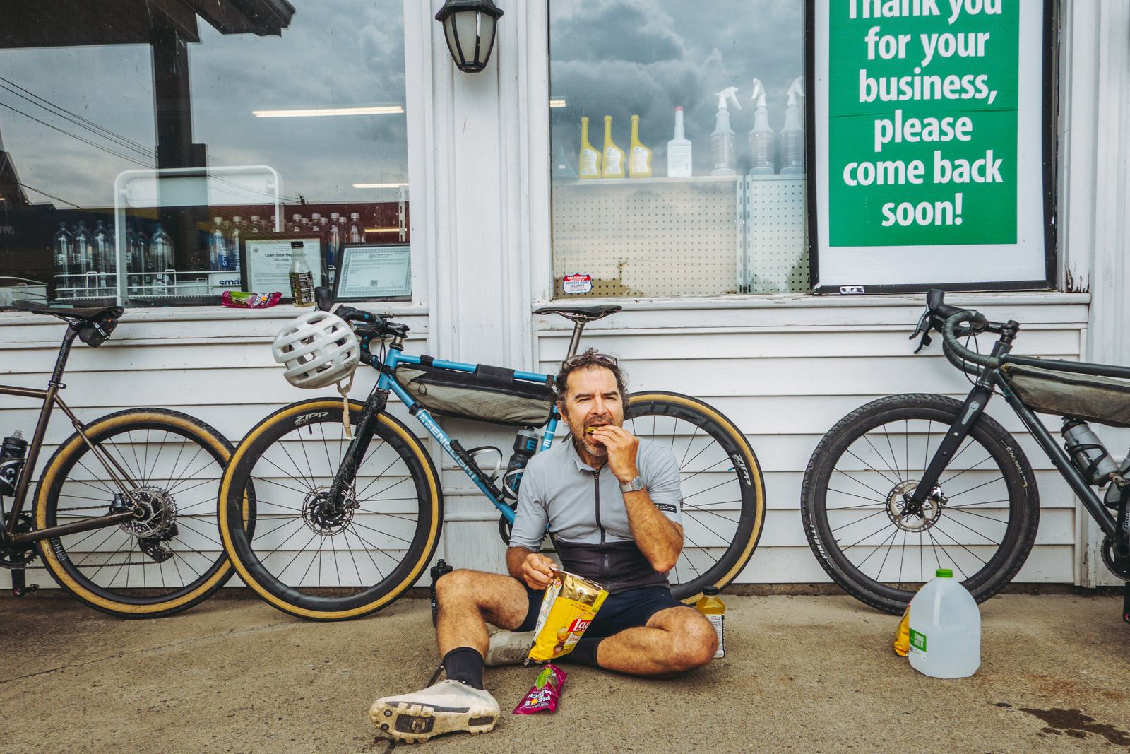A rider sitting on the pavement outside a store eating crisps, bikes propped either side.